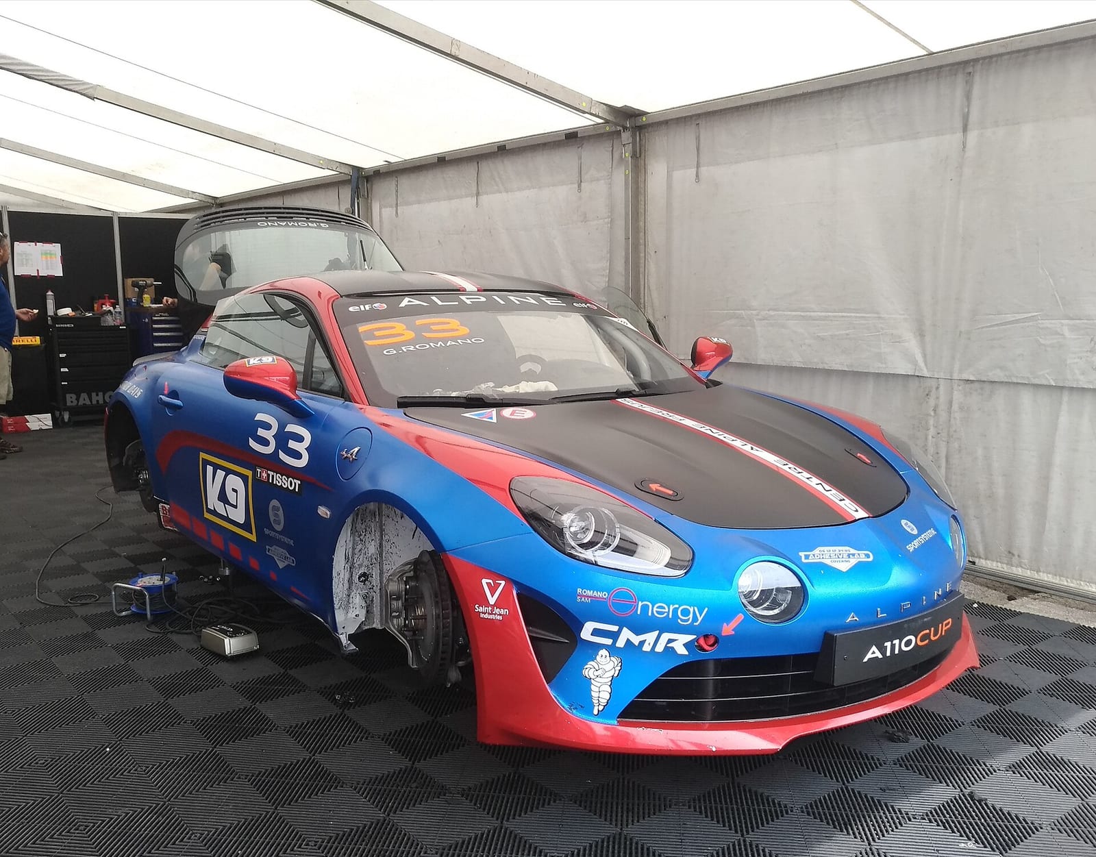 An Alpine A110 Cup race car in blue, white and red in a paddock tent