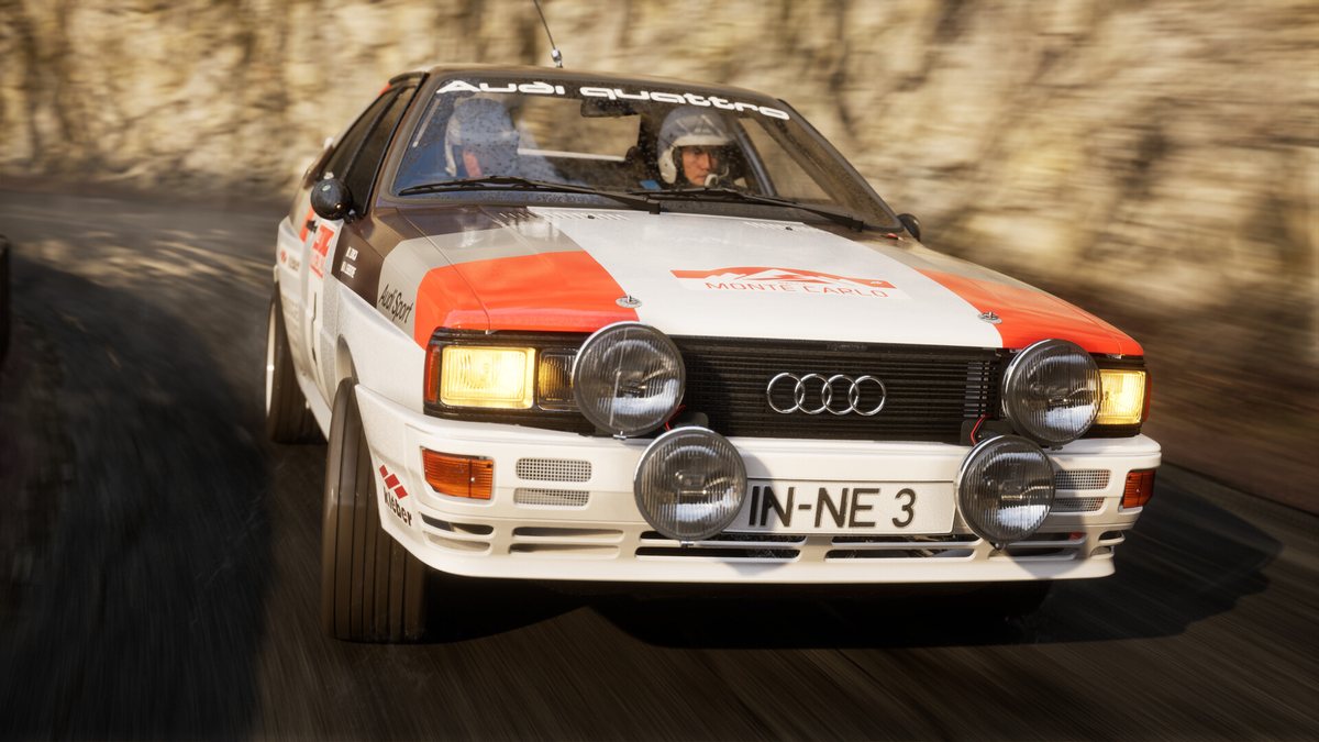 An Audi Quattro Group 4 rally car with its spotlights on, driving on a gravel stage in Assetto Corsa Rally