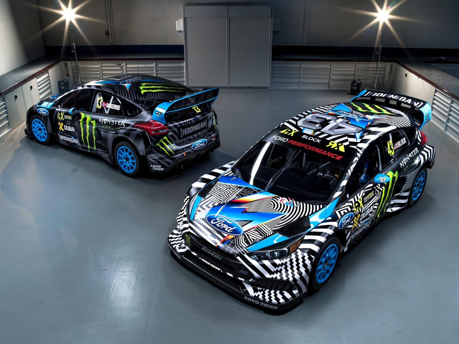 Two Ford Focus RS RX rallycross cars with black, white and blue liveries parked in a workshop