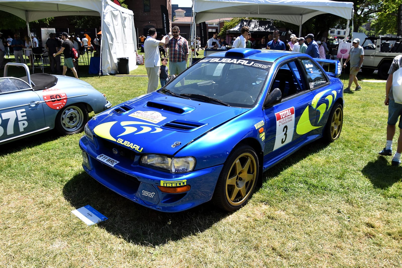 Blue Subaru Impreza WRC from 1998 with gold wheels parked on grass at a car show