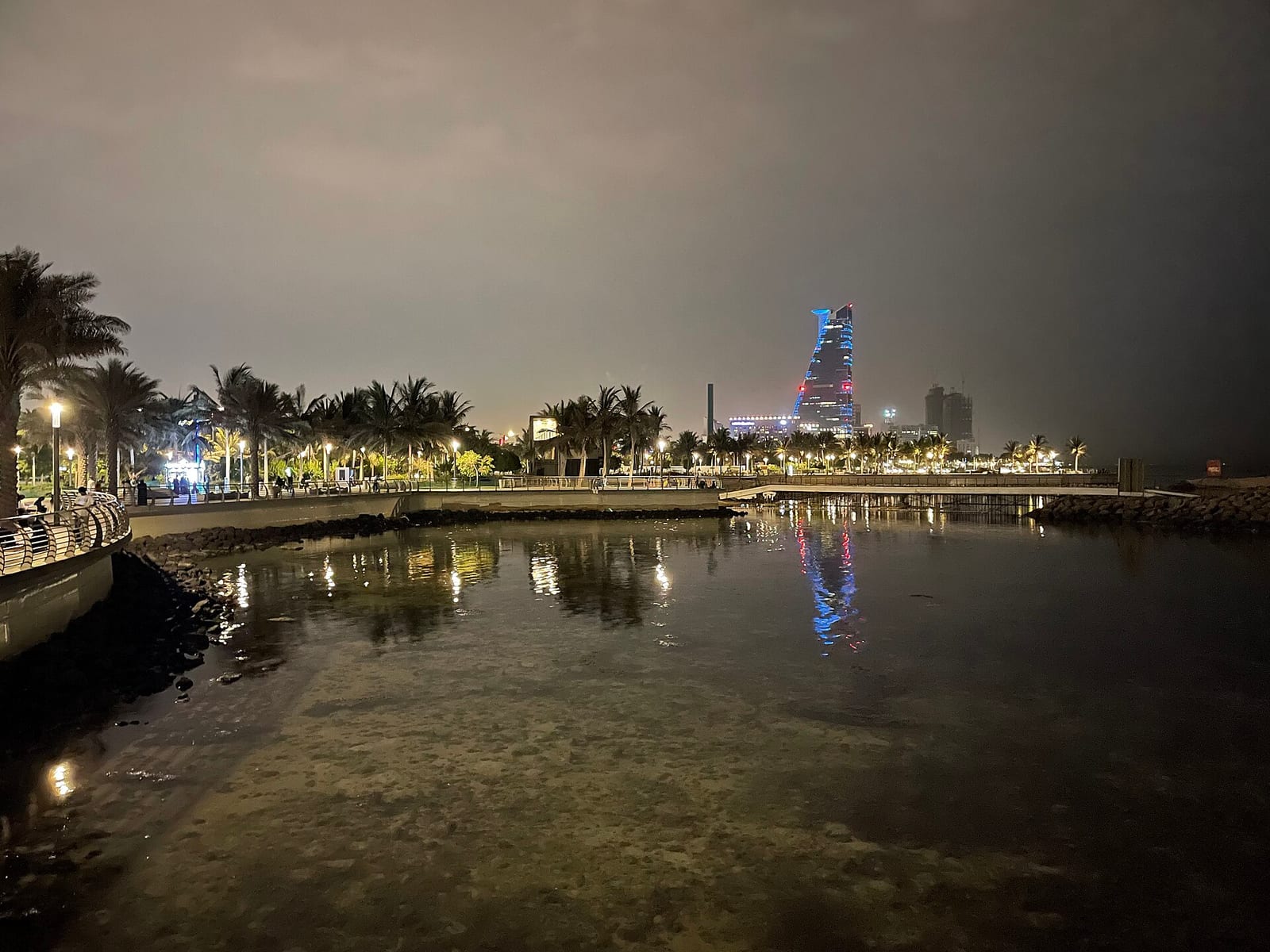 The Jeddah Corniche waterfront at night with palm trees, lights and a lit tower reflected in the water