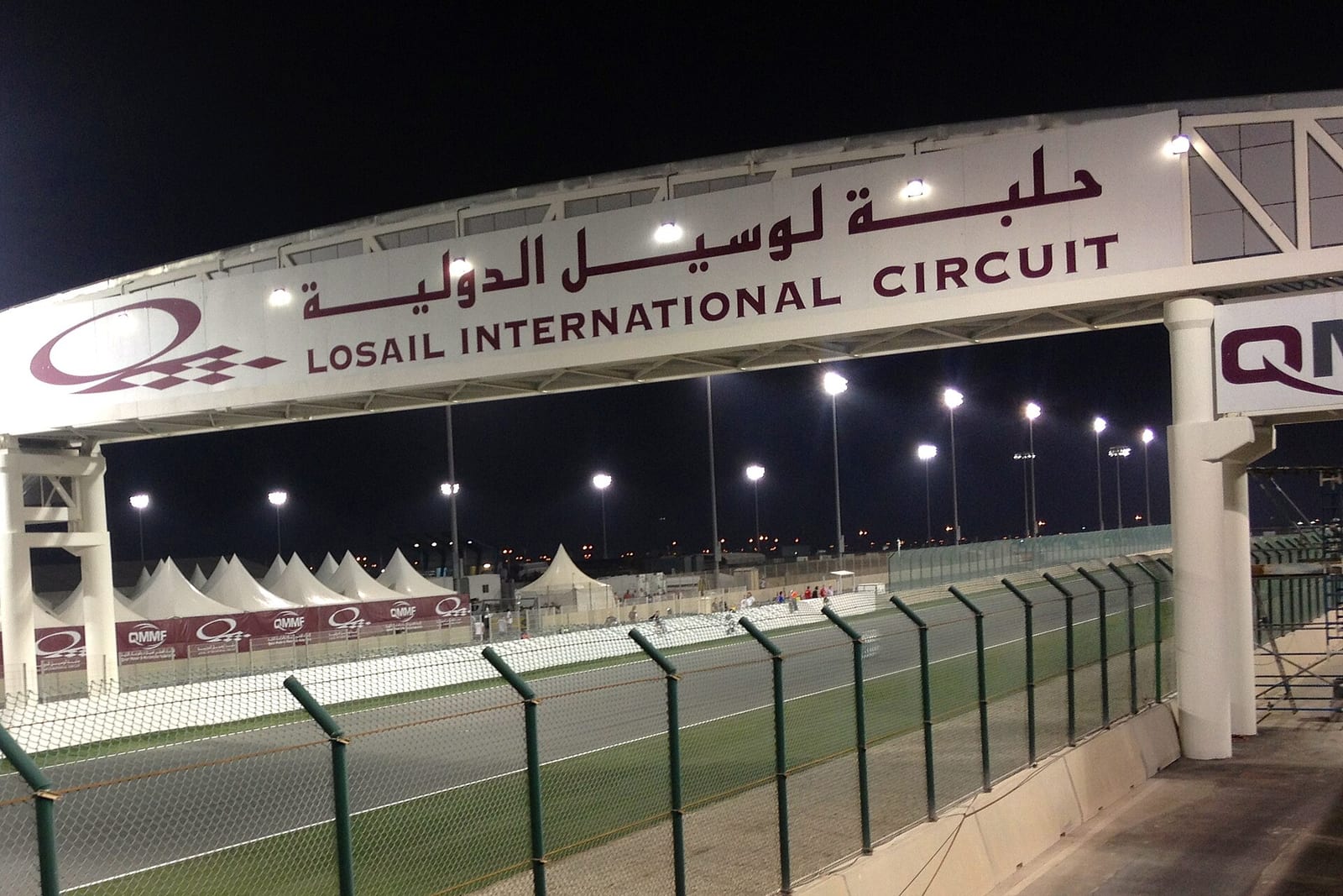 The white entrance board of the circuit at night with the words Losail International Circuit in Arabic and English, and the track behind a fence
