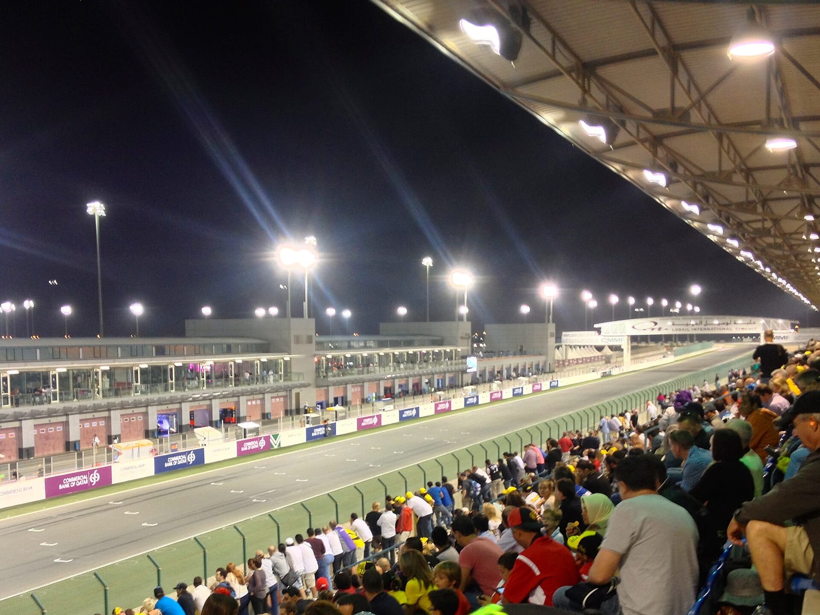 The main straight of the Lusail circuit at night seen from a full grandstand, with the pit lane and the floodlights on the left