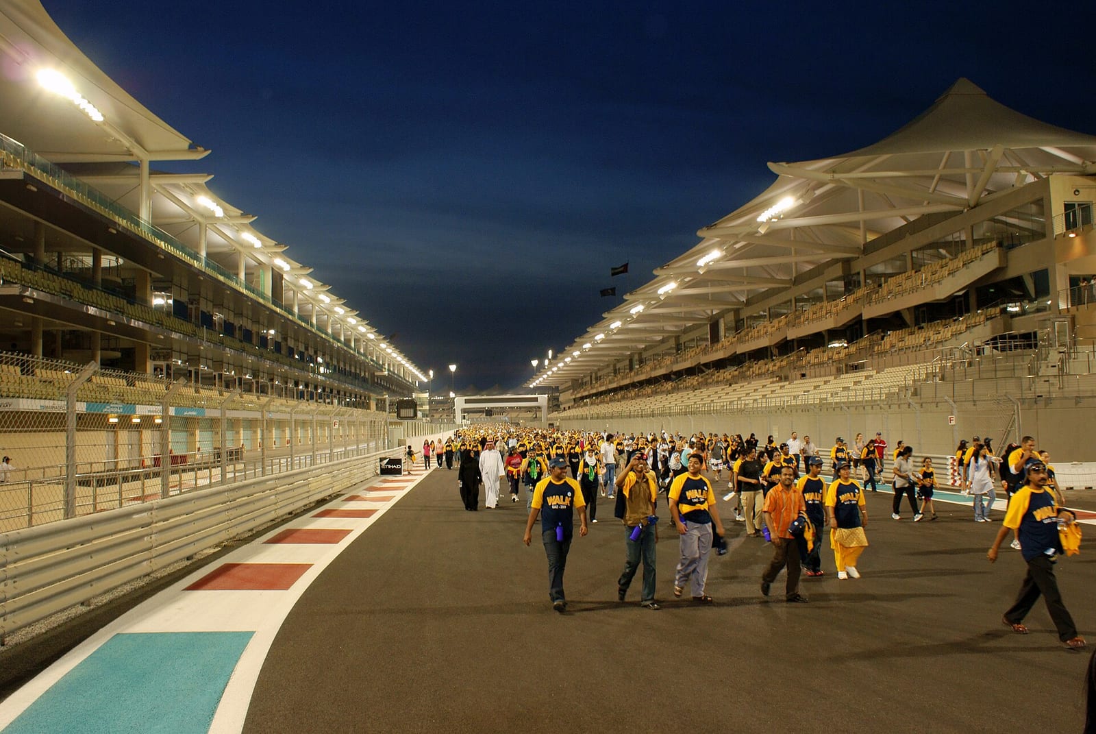 The main straight of the Yas Marina Circuit at night between two lit grandstands, with people walking on the track