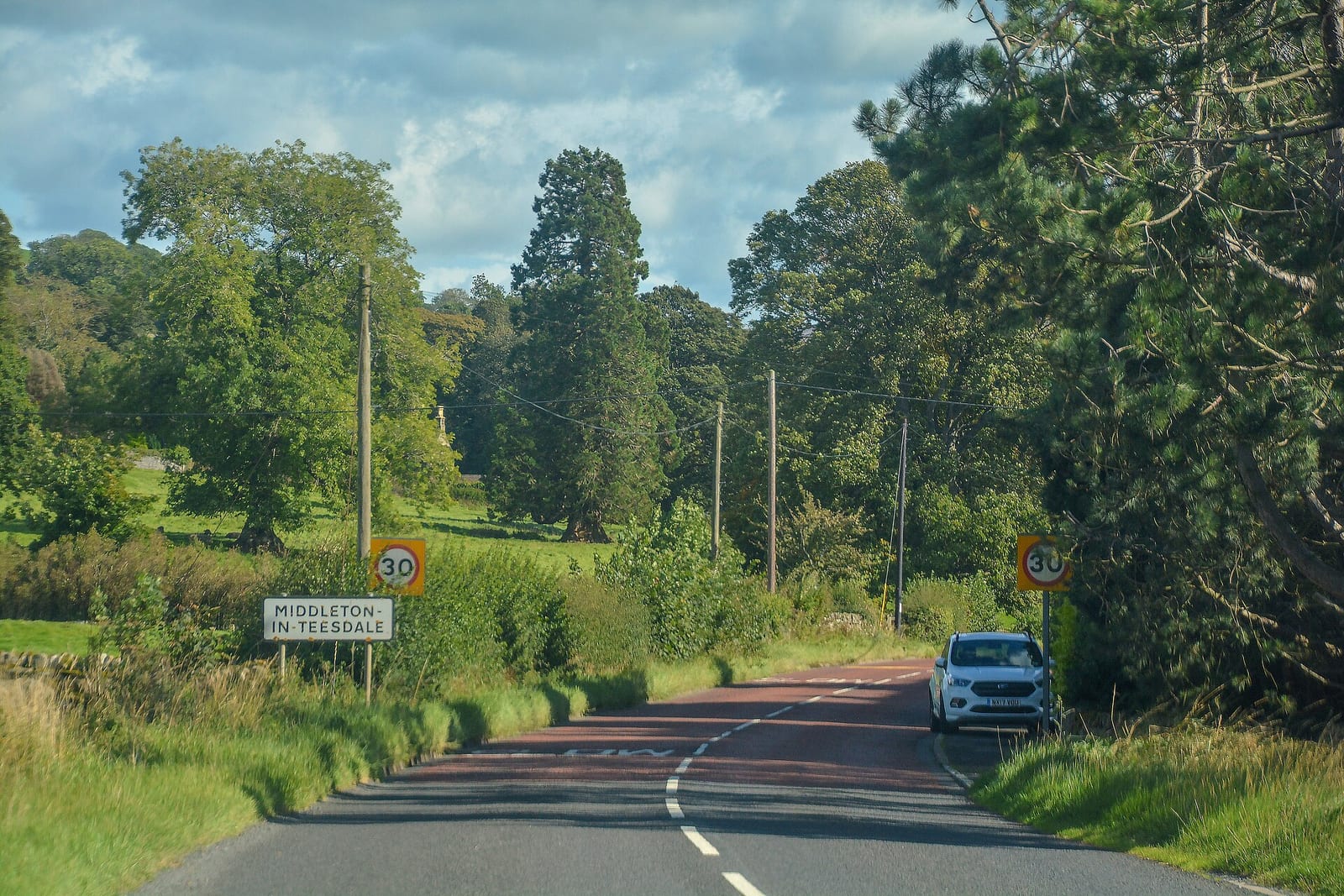 The B6277 road entering Middleton in Teesdale with trees on both sides and a white car parked on the right