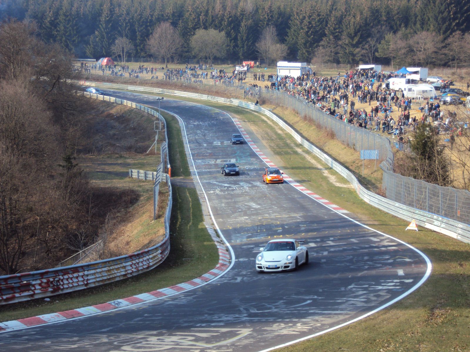 Traffic on the Nordschleife at Brunnchen with a white Porsche 911 GT3 in the foreground and a crowd behind the fence